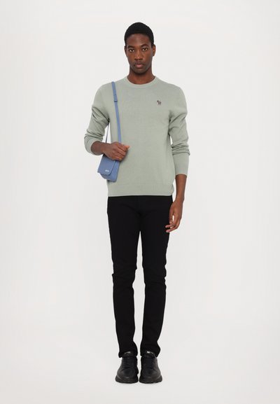 Sweater verde claro, jeans pretos e ténis pretos. Uma pequena mala cruzada azul com um acabamento texturizado é usada atravessada no corpo.