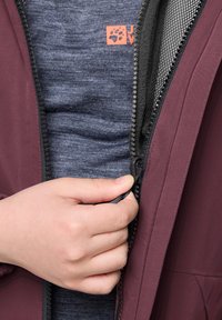 Hand pulling up the zipper on a maroon jacket worn over a blue shirt with an orange logo.