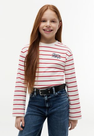 Usmievavé dievča s dlhými červenými vlasmi v bielej dlhoruká blúzka s červenými pruhmi a vyšívaným logom "Tommy Hilfiger", modré džínsy a čierny opasok.