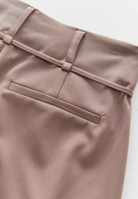 Gros plan d'un pantalon couleur taupe montrant une poche passepoilée horizontale arrière et des passants de ceinture avec une ceinture en tissu assortie.