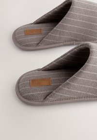 Chaussons en tissu gris avec un motif rayé, une texture douce, un design à dos ouvert et un accent d'étiquette en cuir. Adaptables et confortables.