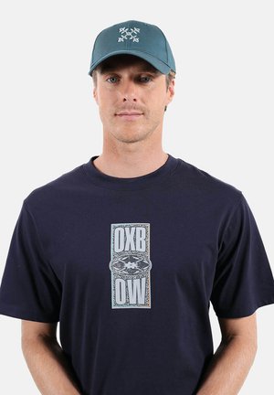 Homme portant une casquette bleue avec un logo de pagaies croisées et un t-shirt bleu marine avec un logo vertical central OXBOW sur un fond uni.