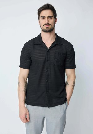Chemise noire à manches courtes avec un motif texturé et un détail de poche en dentelle, présentant un col et une fermeture à boutons.