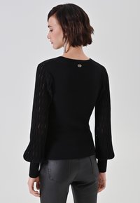 Schwarzer Pullover mit langen, durchsichtigen, gestreiften Ärmeln und einem taillierten Körper. Verfügt über ein kleines Logo am Rückenausschnitt. Kombiniert mit schwarzen Hosen.