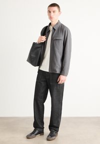 Chaqueta texturizada gris con un bolsillo frontal, camisa crema por debajo, jeans oscuros y zapatillas negras. El modelo lleva un bolso tote negro.