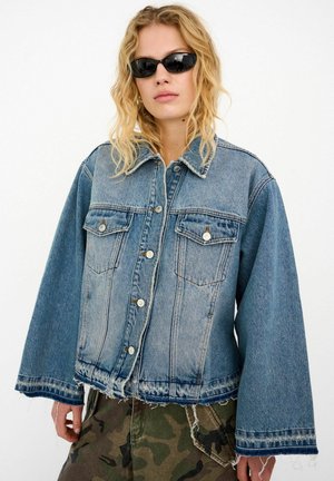 Kurtka jeansowa w jasnoniebieskim kolorze o luźnym kroju, z dwoma kieszeniami na piersi, srebrnymi ozdobami, postrzępionym dołem i szerokimi rękawami, noszona razem z kamuflażowymi spodniami.