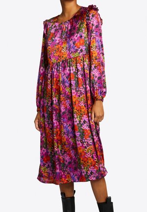 Žena má na sobě dlouhé květované šaty po kolena s růžovými, oranžovými, fialovými a zelenými vzory, doplněné o černé boty.