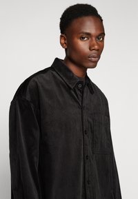 Fekete kordbársony ing nyakkal, gombos záródással, mellzsebbe és függőleges textúrájú csíkokkal. Lazán illeszkedő fazonnal és hosszú ujjakkal rendelkezik.