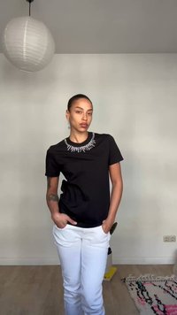 Γυναίκα που στέκεται με τα χέρια στις τσέπες, φορώντας μαύρο πουκάμισο με λευκές διακοσμήσεις και λευκό παντελόνι, σε ένα δωμάτιο με ένα κρεμαστό χαρτοφανάρι.