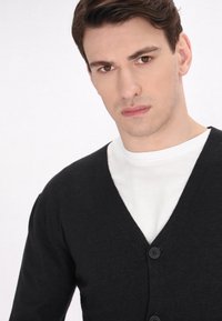 Crna v-neck kardigan od teksturiranog pletenog materijala, s crnim dugmadi, nosi se preko obične bijele majice.
