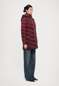 Vrouw in een lange, bordeauxrode pufferjas met capuchon, blauwe wijde jeans en zwarte puntige schoenen, staand in zijaanzicht op een witte achtergrond.