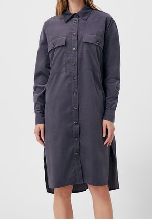 Femme portant une robe-chemise gris foncé à manches longues avec boutons sur le devant et poches poitrine, debout devant un fond clair uni.