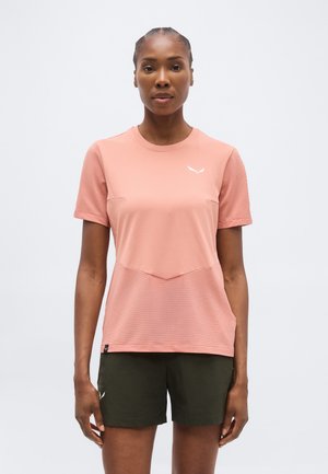 Γυναίκα που στέκεται με το πρόσωπο προς τα εμπρός, φοράει ροζ αθλητική μπλούζα με κοντά μανίκια και σκούρα πράσινα σορτς με απλό λευκό φόντο.