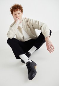 Ljusbeige skjortjacka över en vit skjorta, svarta byxor och svarta sneakers med guldaccent, sitter med en hand på sin haka.