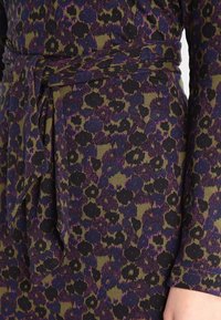 Sukienka w lamparta w ciemnym purpurze, zieleni i czerni. Posiada związany w pasie krój oraz miękką teksturę z dopasowanym fasonem. Detal z długimi rękawami.