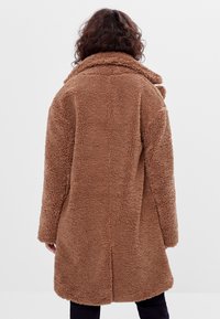 Brun fårskinnsjacka med krage, texturerad tyg, långa ärmar och en loose fit, sedd från baksidan. Inga synliga knäppningar eller mönster.