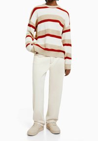 Randig stickad tröja i krämfärg, beige och röd; lös passform med ribbad mudd, i kombination med vita byxor och beige sneakers.
