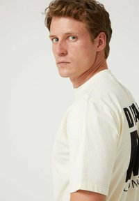 Camiseta de manga corta color crema con texto negro audaz en la espalda, con un ajuste clásico y textura suave de algodón.