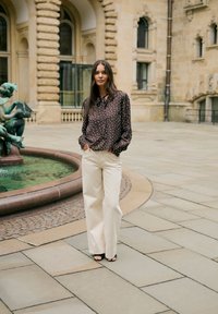 Femme en pantalon beige à jambes larges et blouse sombre à pois, debout près d'une fontaine ronde avec statue dans une cour en pierre.