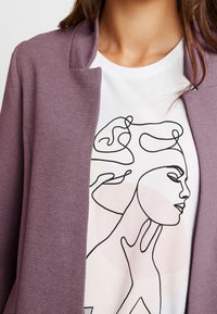 Gros plan d'une personne portant une chemise blanche avec un dessin abstrait en ligne continue représentant un visage et une main de femme, ainsi qu'une veste texturée violette.