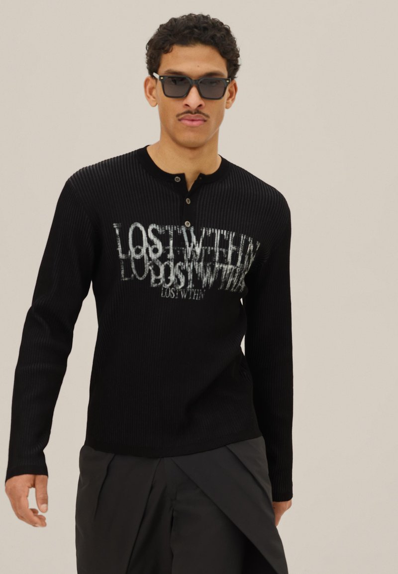 Homme portant un t-shirt noir à manches longues côtelé avec le texte « LOST WITHIN », des lunettes de soleil noires et un pantalon foncé, sur un fond beige uni.
