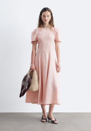 Frau mit langen Haaren, die ein blassrosa Midikleid trägt, hält eine geflochtene Tasche mit einem dunkel gemusterten Schal und trägt schwarze offene High Heels.