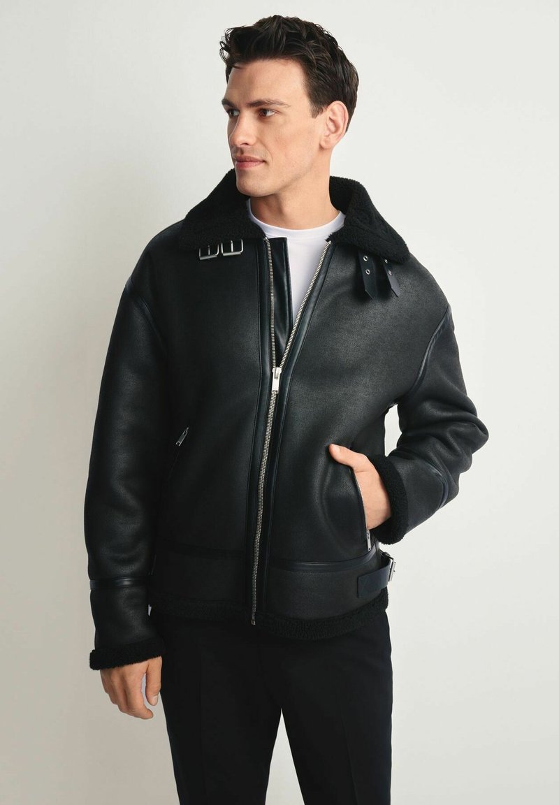Czarna skórzana kurtka z kołnierzem z owczej wełny, zapinana na zamek, z bocznymi kieszeniami i akcentami w postaci klamr; charakteryzuje się gładką teksturą i solidnym wykonaniem.