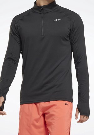 Mies pukeutuneena mustaan pitkähihamaiseen puolivetoketjulliseen urheilupaitaan ja korallinvärisiin shortseihin, joissa on peukaloreiät hihoissa ja brändilogot rinnassa ja shortseissa.