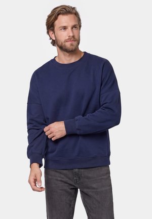 Mand med skæg iført en mørkeblå sweatshirt og mørke jeans, der justerer venstre ærme, med en neutral lysegrå baggrund.