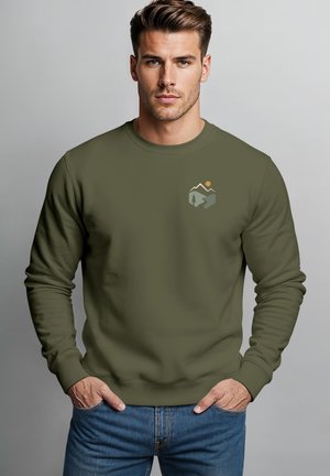 Mann mit kurzen braunen Haaren trägt einen olivgrünen Pullover mit Berg-, Baum- und Sonnenmotiv und steht mit den Händen in den Taschen der blauen Jeans.