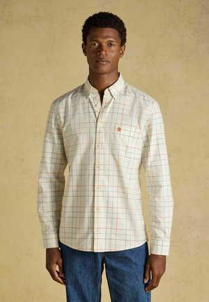 Mann trägt ein cremefarbenes langärmeliges Hemd mit orange-blauem Karomuster und blaue Jeans, steht vor beigem Hintergrund.