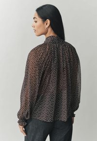 Kerge must blouse valgete täppidega, volditud kaelus ja elastsed mansetid, koos tumedate teksadega.