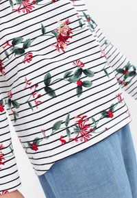 Witte gestreepte shirt met rode bloemenprint, voorzien van groene bladeren. Het shirt heeft lange mouwen en een afgeronde zoom, gecombineerd met blauwe broek.