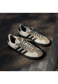 Bež i bijele tenisice s tri crne pruge, teksturiranom gornjom površinom, sivim naglascima i gumenim đonom, dizajnirane za svakodnevno nošenje.