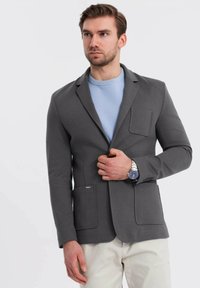 Hombre con blazer gris sobre suéter azul claro y pantalones beige, ajustándose el blazer con la mano izquierda, lleva un reloj de plata con carátula oscura.