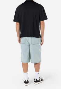 Man die een zwart T-shirt met korte mouwen, lichtblauwe spijkershorts, witte sokken en zwart-witte sneakers draagt, staand met de rug naar de camera gekeerd.