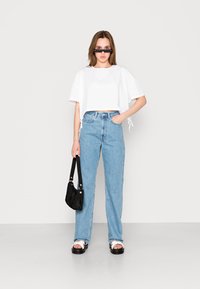 T-shirt branca cropped, jeans de perna reta azul claro, mala mini preta e sandálias chunky pretas com correias brancas. A modelo usa óculos de sol estreitos.
