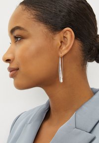 Des boucles d'oreilles en argent, allongées en forme de créoles, pendent de l'oreille. Le design élégant présente une surface polie et réfléchissante avec une texture lisse.
