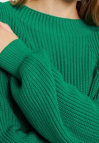 Zelený pletený svetr s žebrovanou texturou, širokým výstřihem a volnými rukávy. Tkanina působí měkce a hustě.