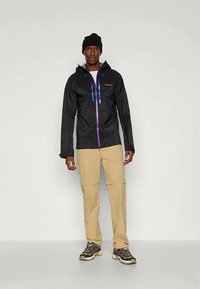 Schwarze wasserdichte Jacke mit lila Reißverschlüssen, einer Kapuze und verstellbaren Schnüren, kombiniert mit beigen Hosen und dunklen Schuhen.