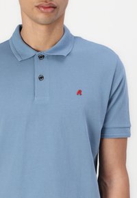 Svetlo modra polo majica iz teksturirane tkanine s ovratnikom, tremi črnimi gumbi in majhnim rdečim logotipom na levi prsni strani.