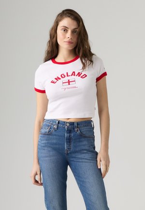 Mladá žena s dlhými hnedými vlasmi, oblečená v bielom krátkom tričku s červeným lemom a nápisom "England" a modrých vysokých džínsach.