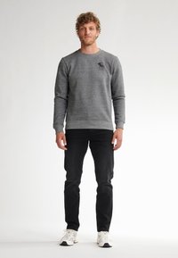 Grijze sweatshirt met een ronde hals, geribbelde mouwen en een klein logo op de borst, gecombineerd met zwarte jeans en witte sneakers.