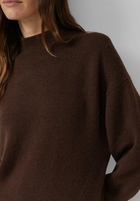 Brauner Strickpullover mit hohem Kragen, überschnittenen Schultern und weicher, strukturierter Oberfläche. Details sind gerippte Bündchen an Ärmeln und Saum.