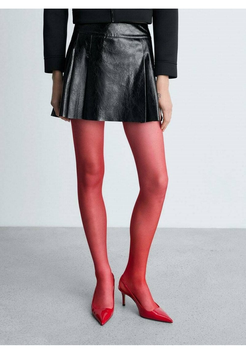 Personne portant une mini-jupe en cuir noire, des collants rouges transparents et des talons hauts rouges à bout pointu, debout sur un sol gris clair.