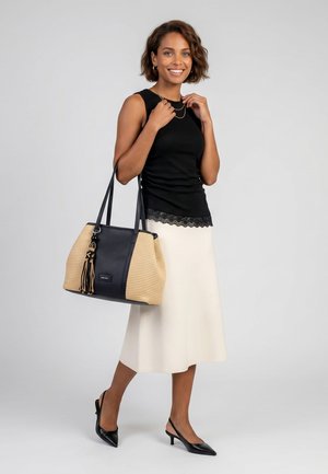 Donna sorridente, indossa un top nero senza maniche e una gonna crema, tiene una grande borsa beige e nera, tacchi neri slingback, in piedi davanti a uno sfondo grigio.