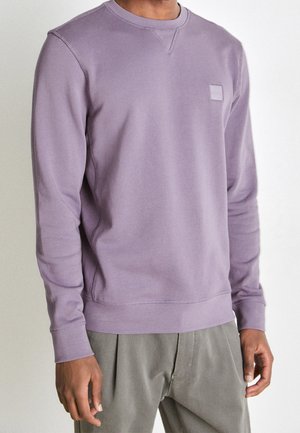 Mann trägt einen lavendelfarbenen Langarm-Crewneck-Pullover mit kleinem rechteckigem Logoaufnäher auf der Brust und graue Hose.