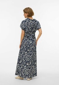Femme aux cheveux courts vue de dos, portant une robe maxi à motifs bleu marine et blanc nouée à la taille, ainsi que des chaussures plates beiges perforées.