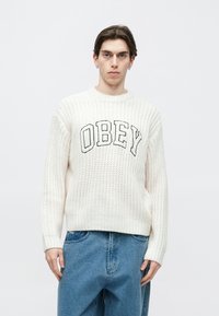 Pulover alb tricotat cu logo negru „OBEY” pe piept, cu guler rotund și manșete ribbed, asortat cu pantaloni din denim albastru.