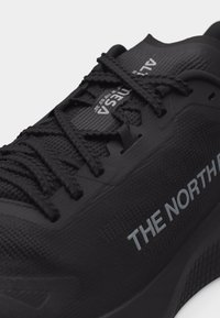Chaussure de sport noire avec tissu texturé, lacets noirs et logo "THE NORTH FACE" sur le côté.